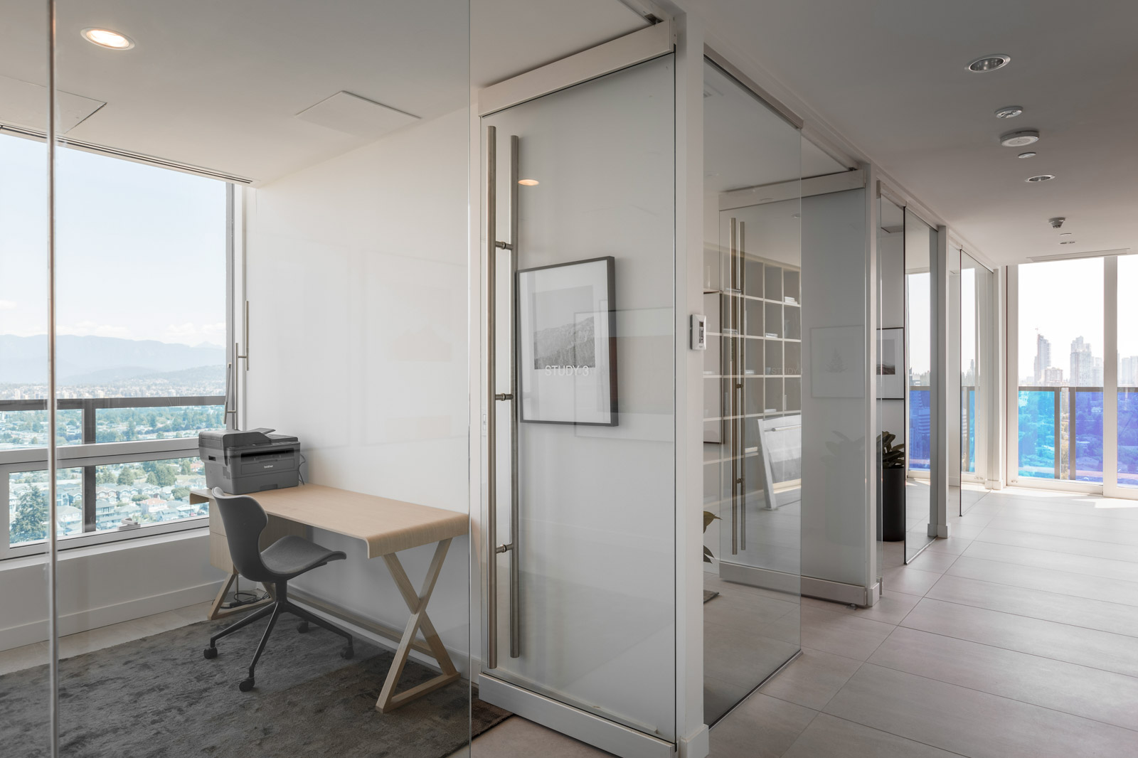 Modern office space with glass walls, a desk with a chair and printer, large windows, and a city view in the background.