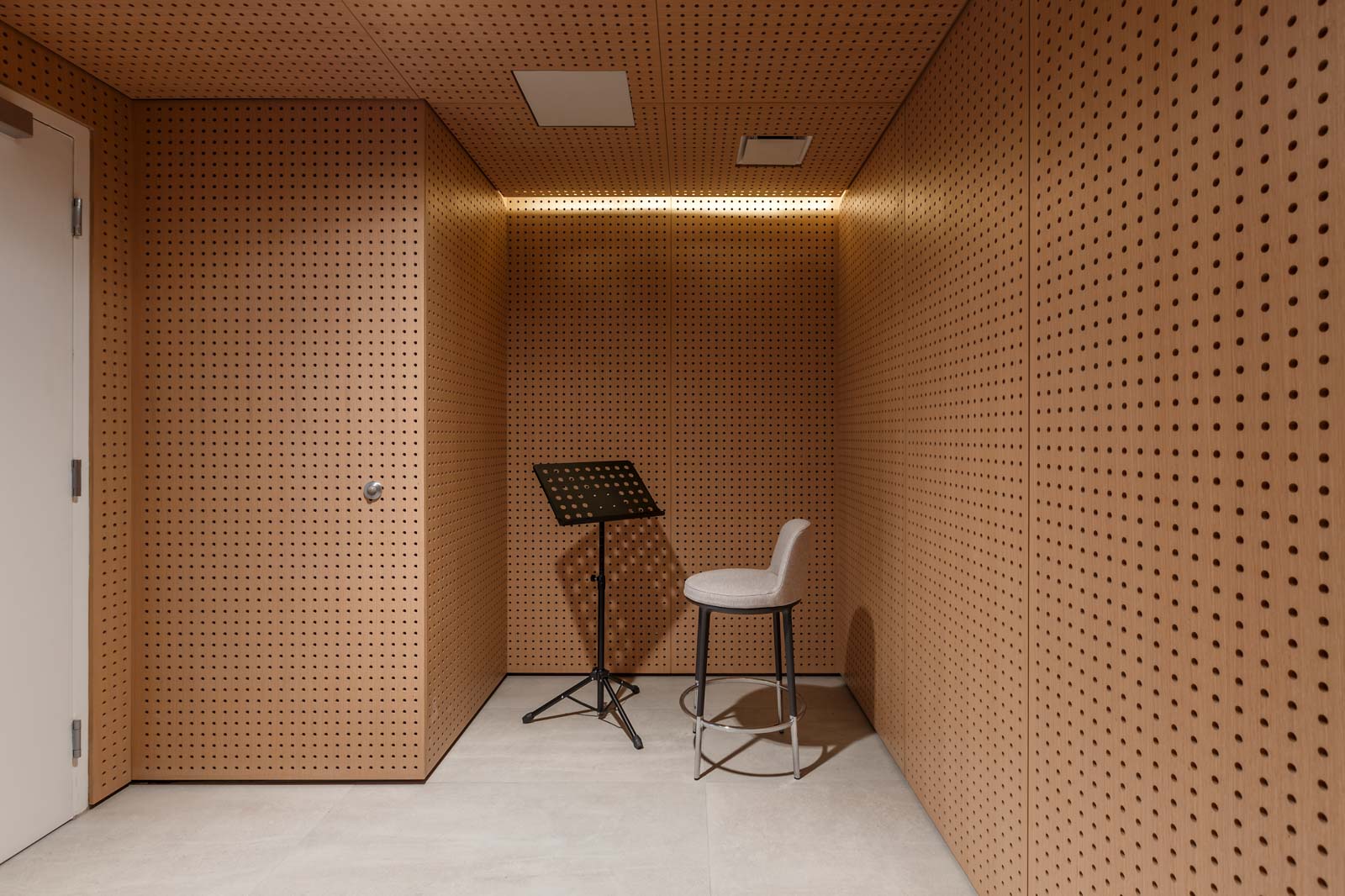 A small soundproof room with perforated wooden panels, a music stand, and a padded stool on a light-colored tiled floor.