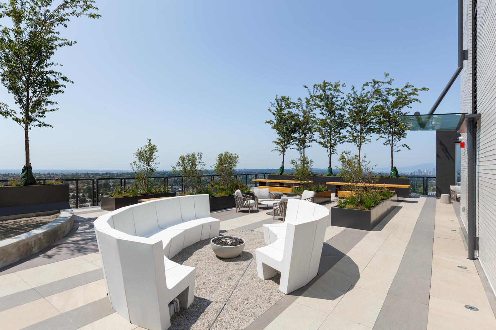 Modern rooftop terrace with white curved seating around a fire pit, planters with trees, a long wooden bench, and a city view in the background.