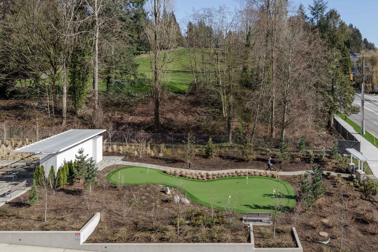 Aerial view of a small outdoor putting green next to a white structure, surrounded by trees and landscaping near a road.