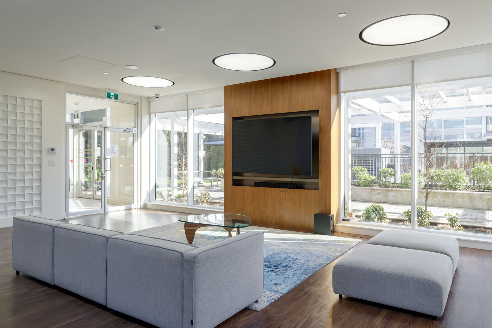 Modern lounge with a large gray sectional sofa, glass coffee table, wall-mounted TV on wood panel, floor-to-ceiling windows, and patio view.