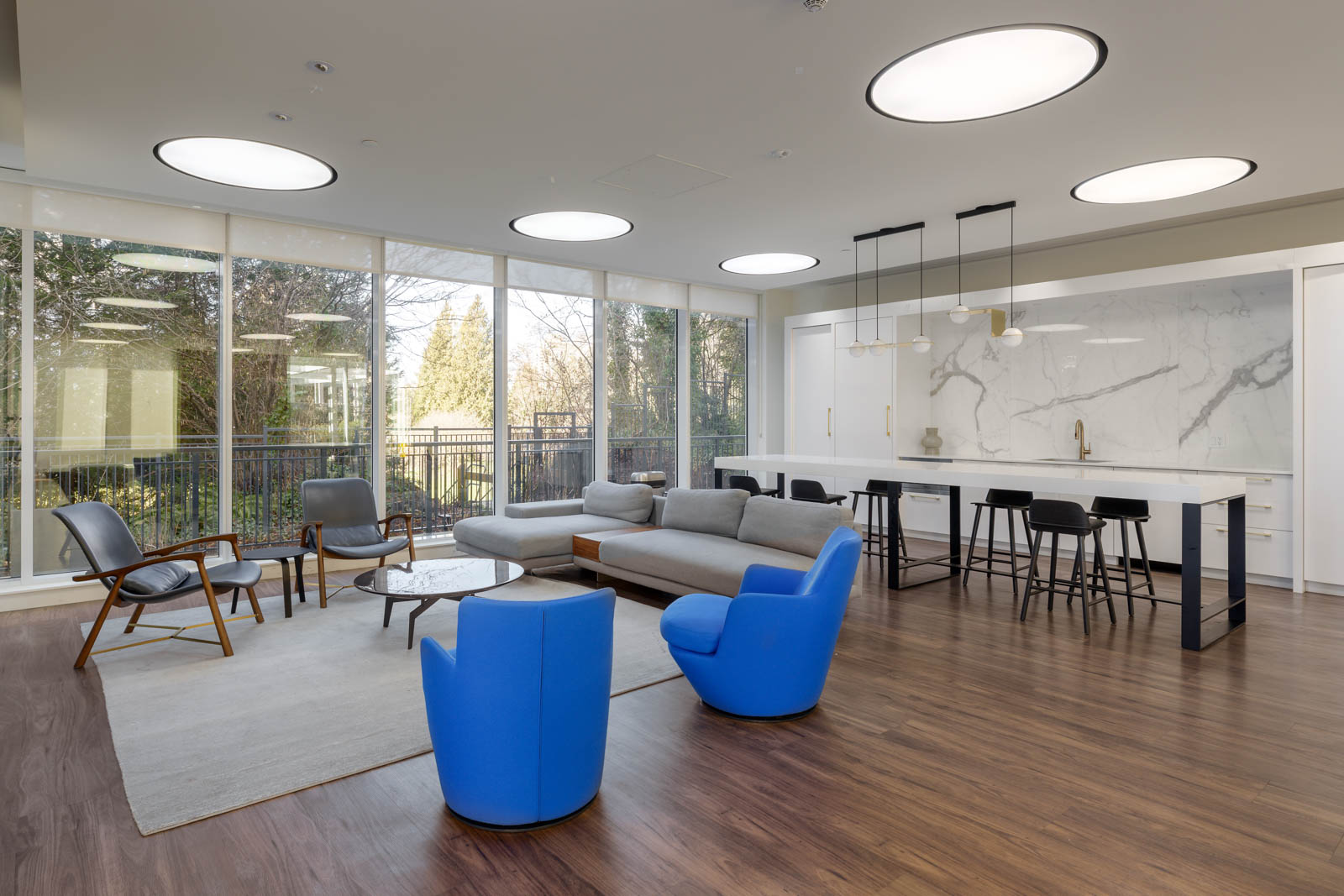 Modern lounge area with blue and gray seating, a coffee table, and a bar with stools; large windows provide natural light and view of trees outside.