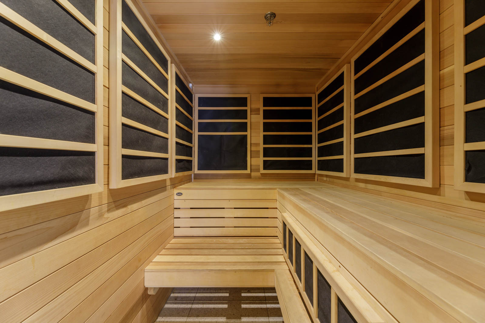 Interior of a modern infrared sauna with wooden benches and walls lined with black infrared heating panels, illuminated by a ceiling light.