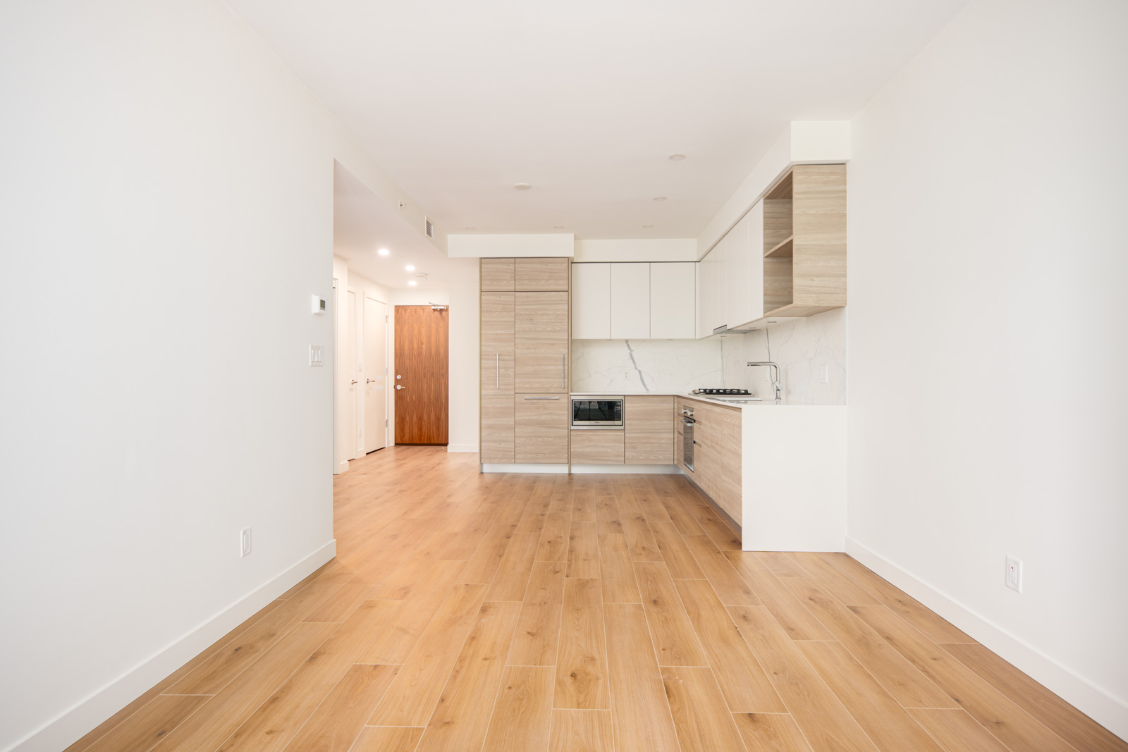 Modern empty apartment with wood flooring, white walls, and a kitchen area with light wood cabinets and built-in appliances. Hallway leads to other rooms.