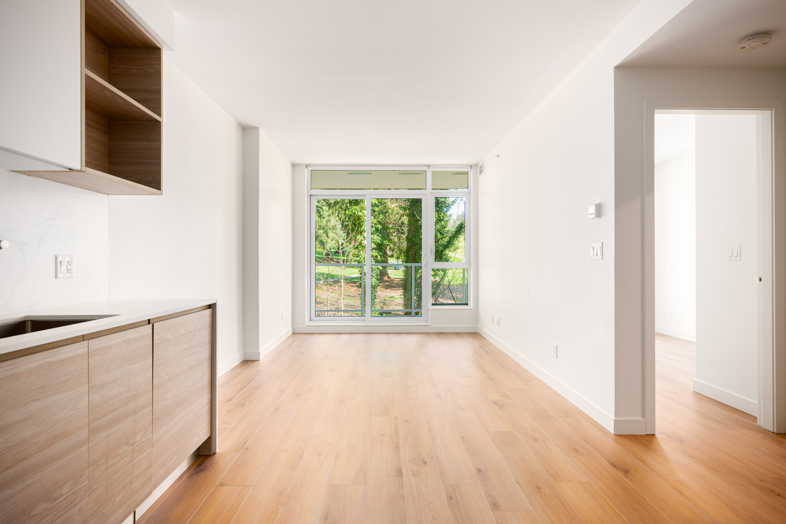A modern, unfurnished apartment with light wood floors, white walls, large window, and an open kitchen area. Sunlight enters from the window, and a doorway leads to another room.