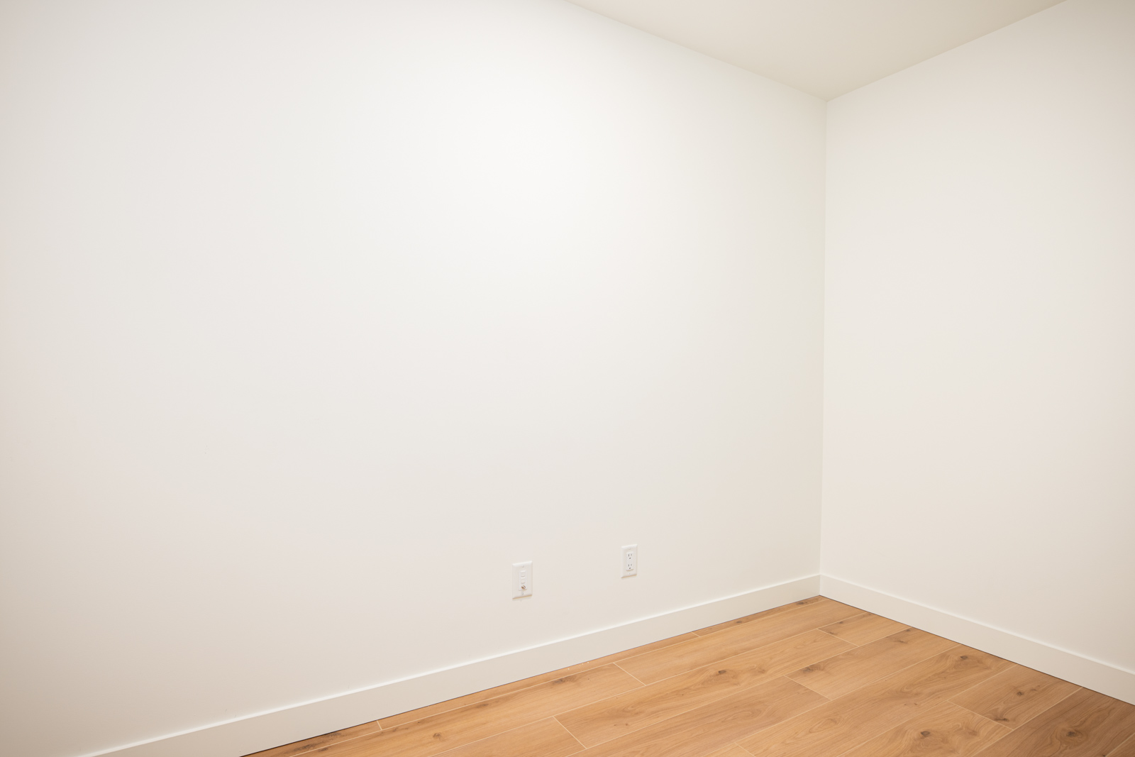 Empty room with white walls, light wood flooring, and two electrical outlets on the lower part of the wall.