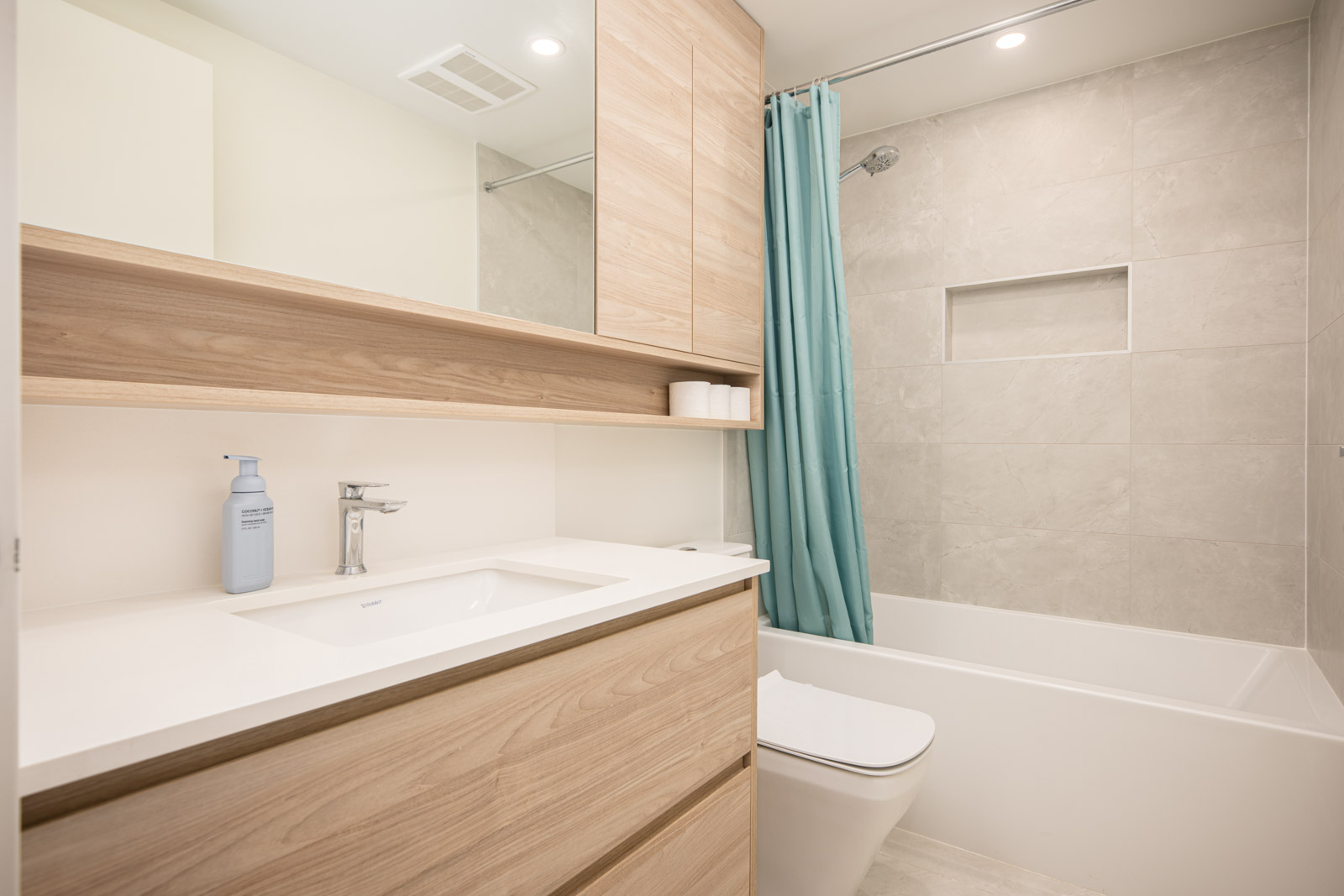 Modern bathroom with light wood cabinets, white countertop, wall-mounted mirror, toilet, bathtub with blue shower curtain, and beige tiled walls.