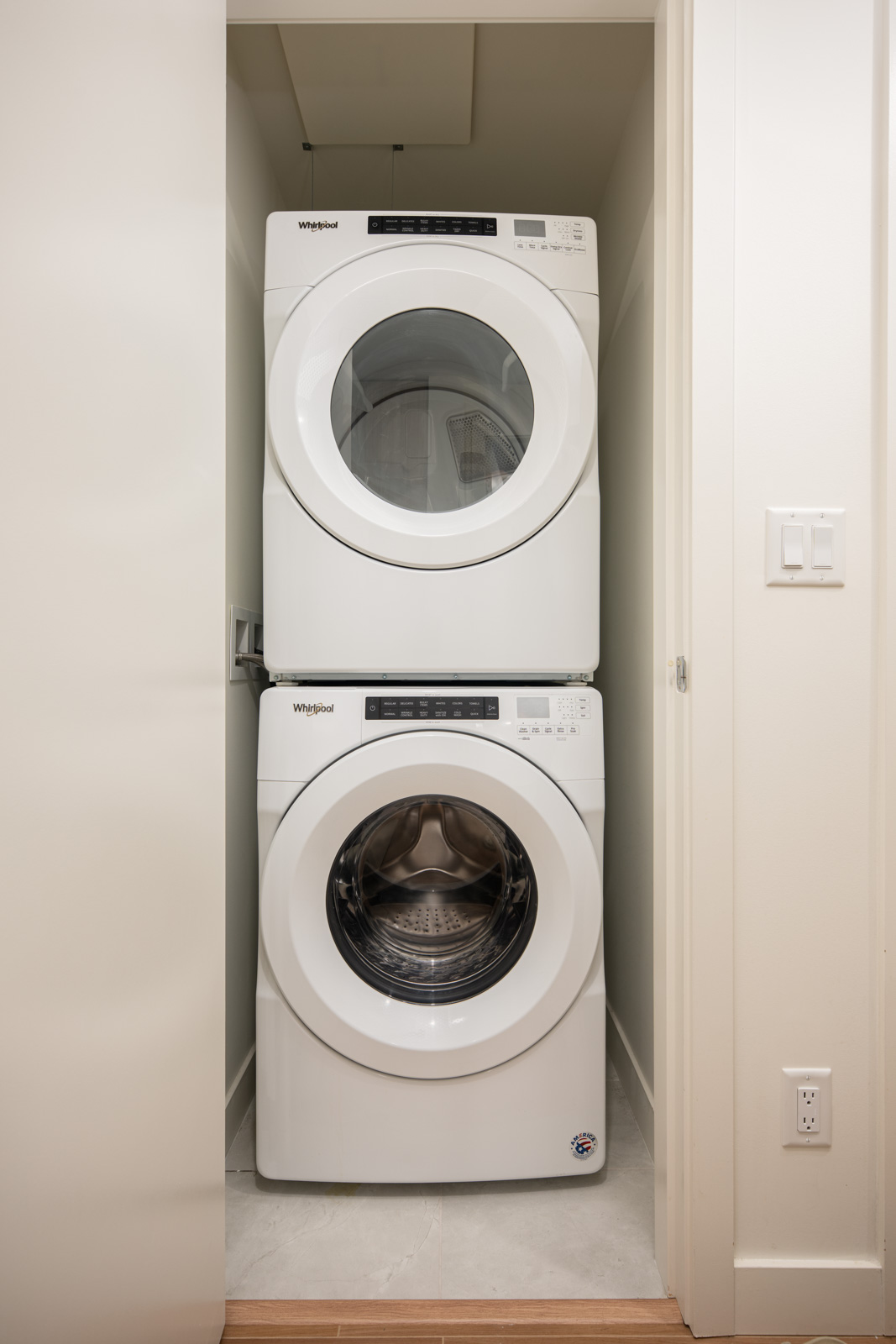 A stacked washer and dryer set is placed in a small, white-walled laundry closet with a closed door and light-colored flooring.