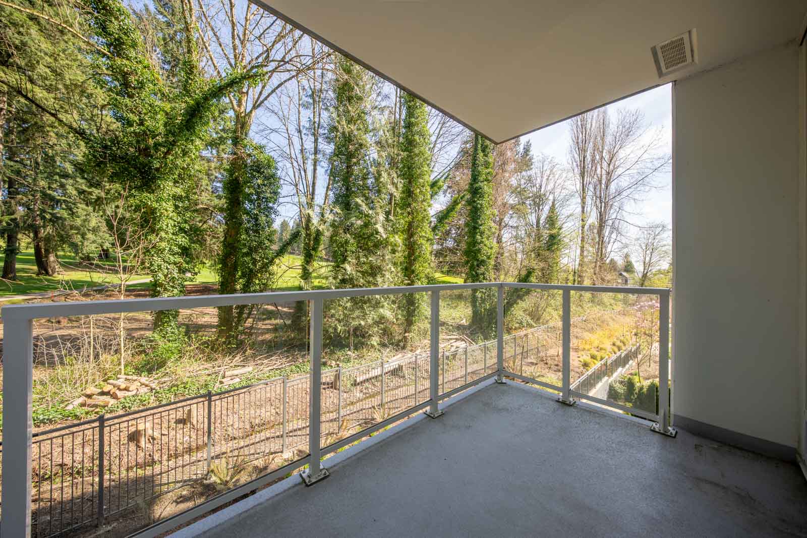 A covered balcony with a glass railing overlooks a fenced yard and tall trees in a wooded outdoor area on a clear day.