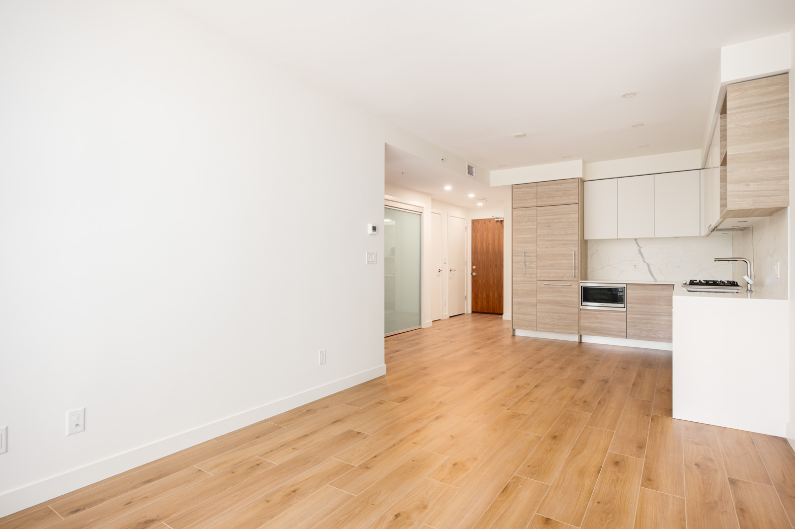 Modern, unfurnished apartment with light wood floors, white walls, a small kitchen area with wood and white cabinets, and a hallway leading to a closed wooden door.