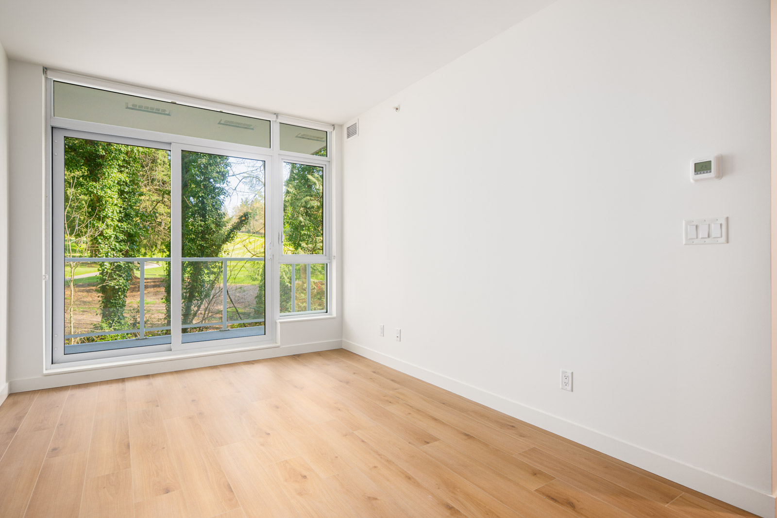 Empty room with light wood flooring, white walls, and large floor-to-ceiling windows overlooking a green outdoor area.