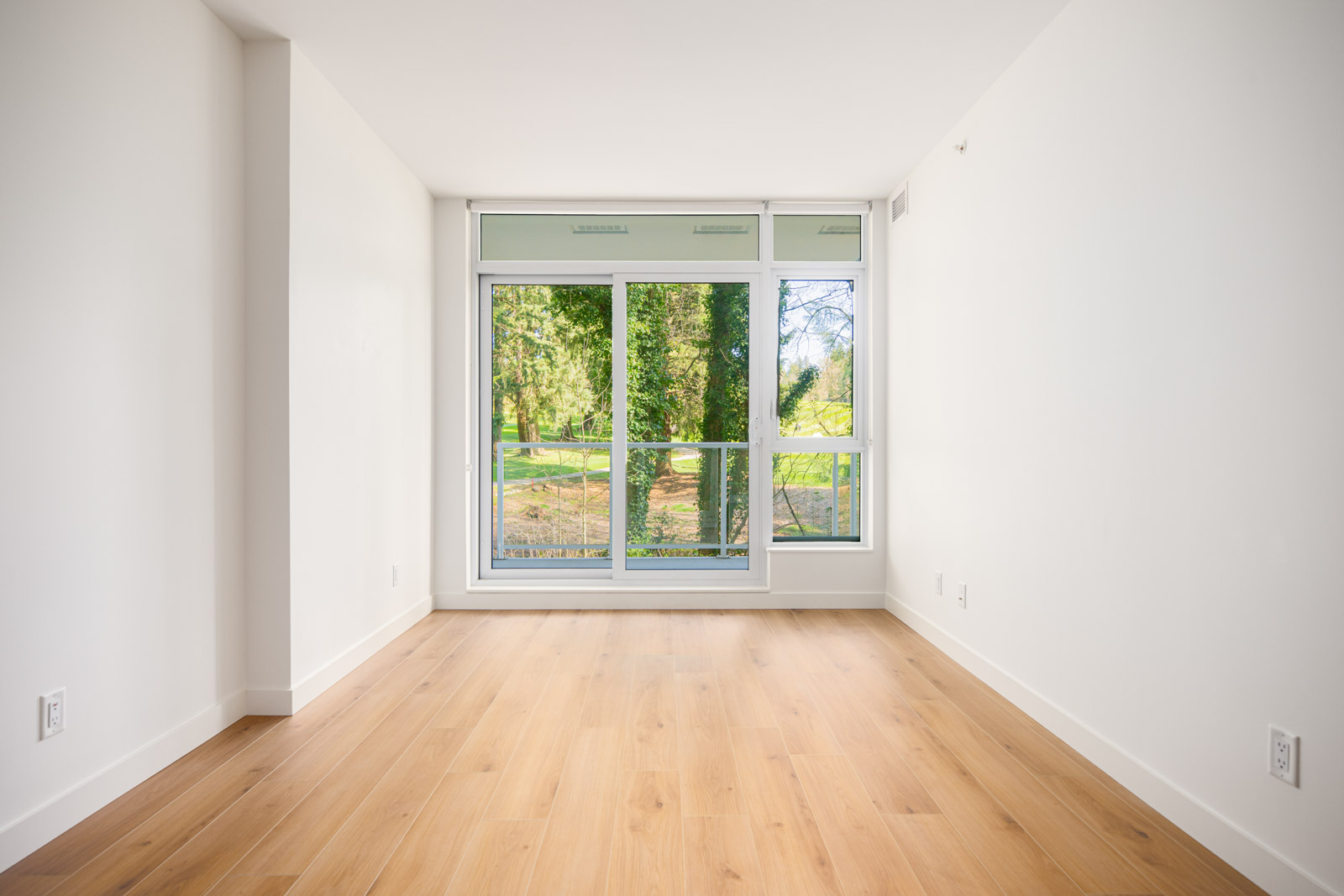 Empty room with light wood flooring, white walls, and a large window overlooking green trees and a grassy outdoor area.