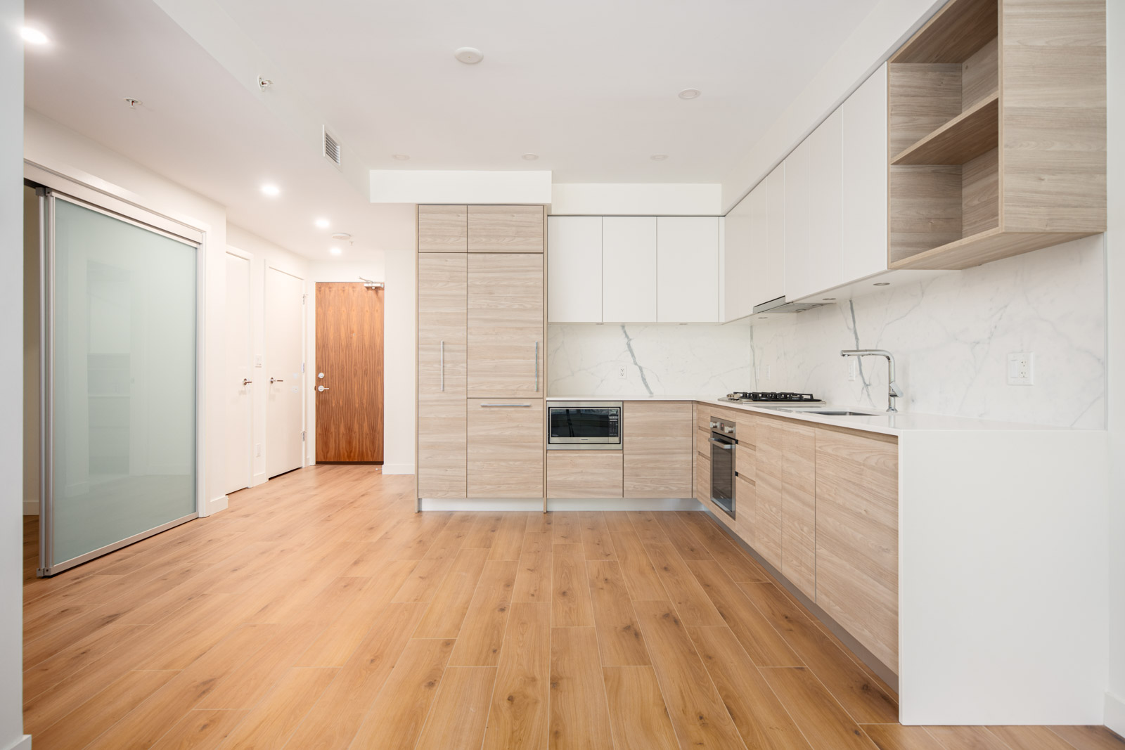 Modern kitchen with light wood cabinets, white countertops, built-in appliances, and marble backsplash; open layout with light wood flooring and sliding glass doors.