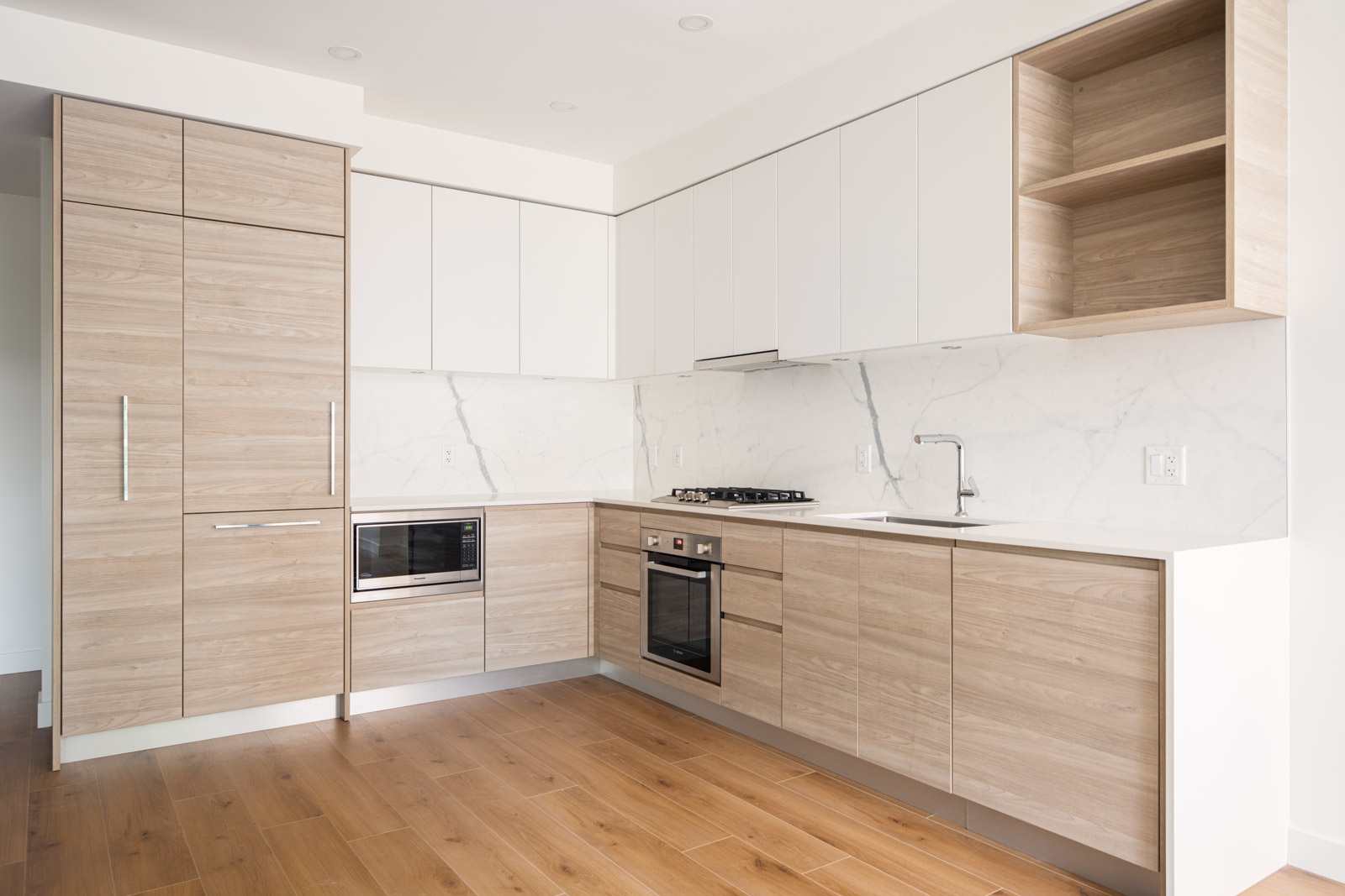 Modern kitchen with light wood cabinets, white upper cabinets, marble backsplash, built-in appliances, stainless steel sink, and wood flooring.