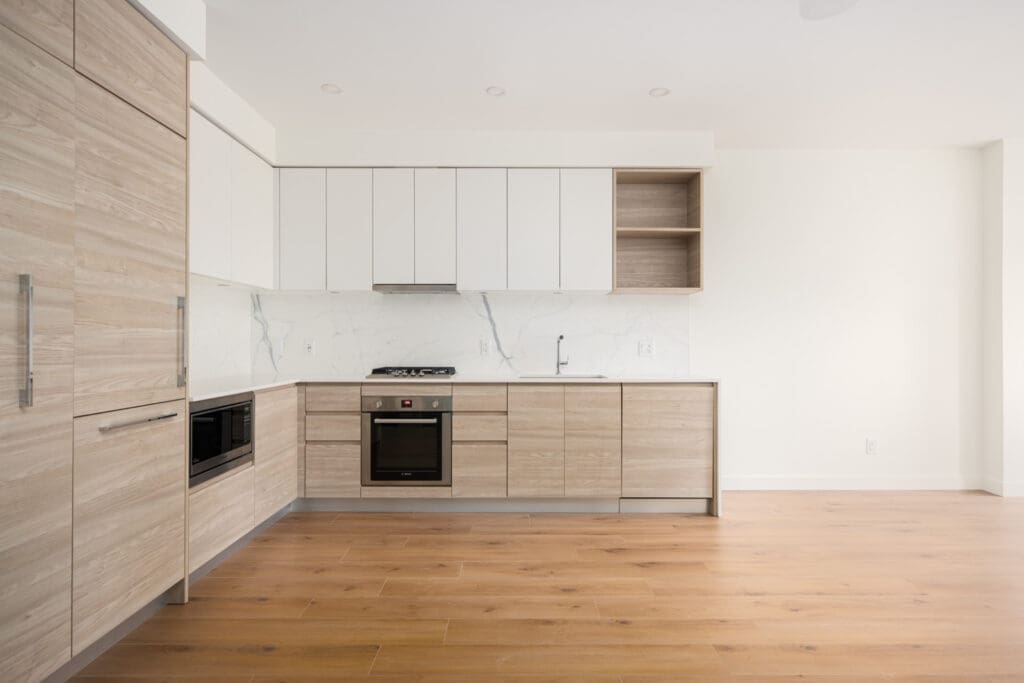 Modern kitchen with light wood cabinets, white upper cabinets, marble backsplash, built-in oven, and hardwood flooring. The space is clean and minimalistic with neutral tones.