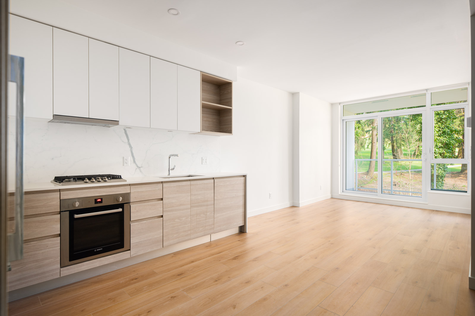 Modern kitchen with light wood cabinets, built-in oven, and large window overlooking a green outdoor area, featuring light wood flooring and white walls.