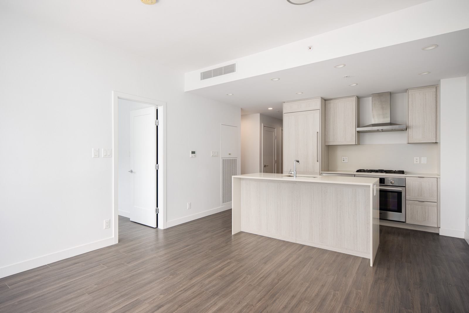 A modern, unfurnished kitchen and living area with light wood cabinetry, island, stainless steel appliances, and wood flooring. White walls and ample natural light.