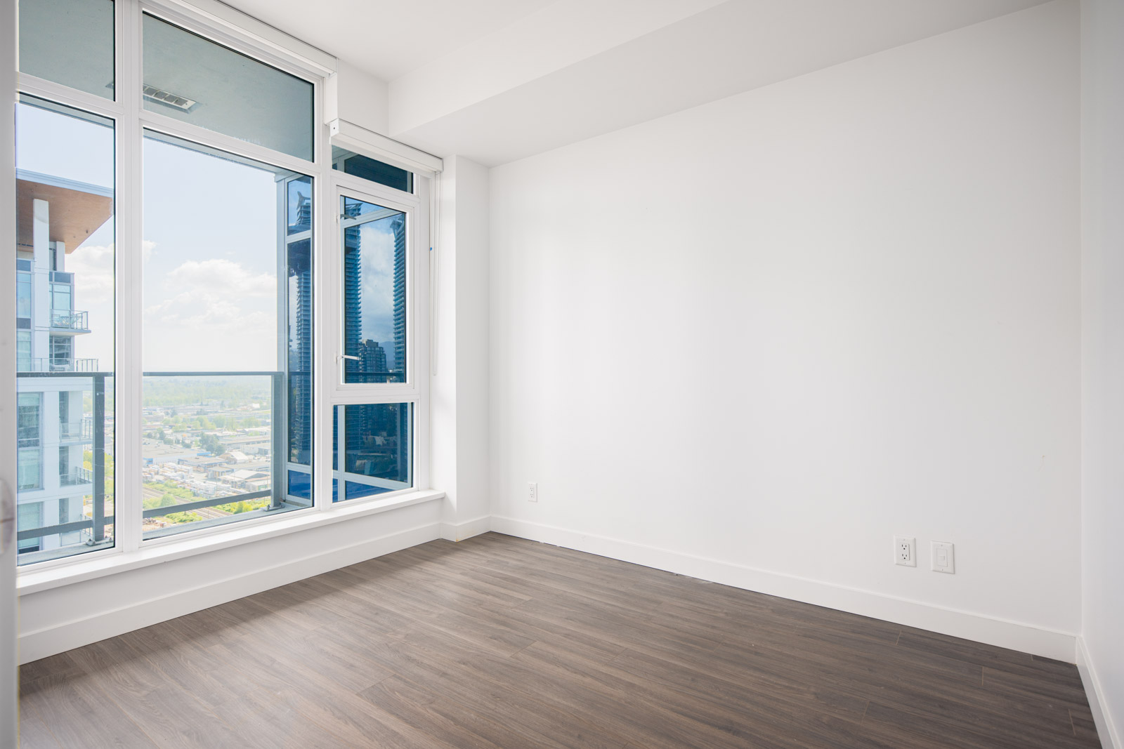 Empty modern room with white walls, large windows, and wood-style flooring, showing a view of nearby buildings and a distant landscape.