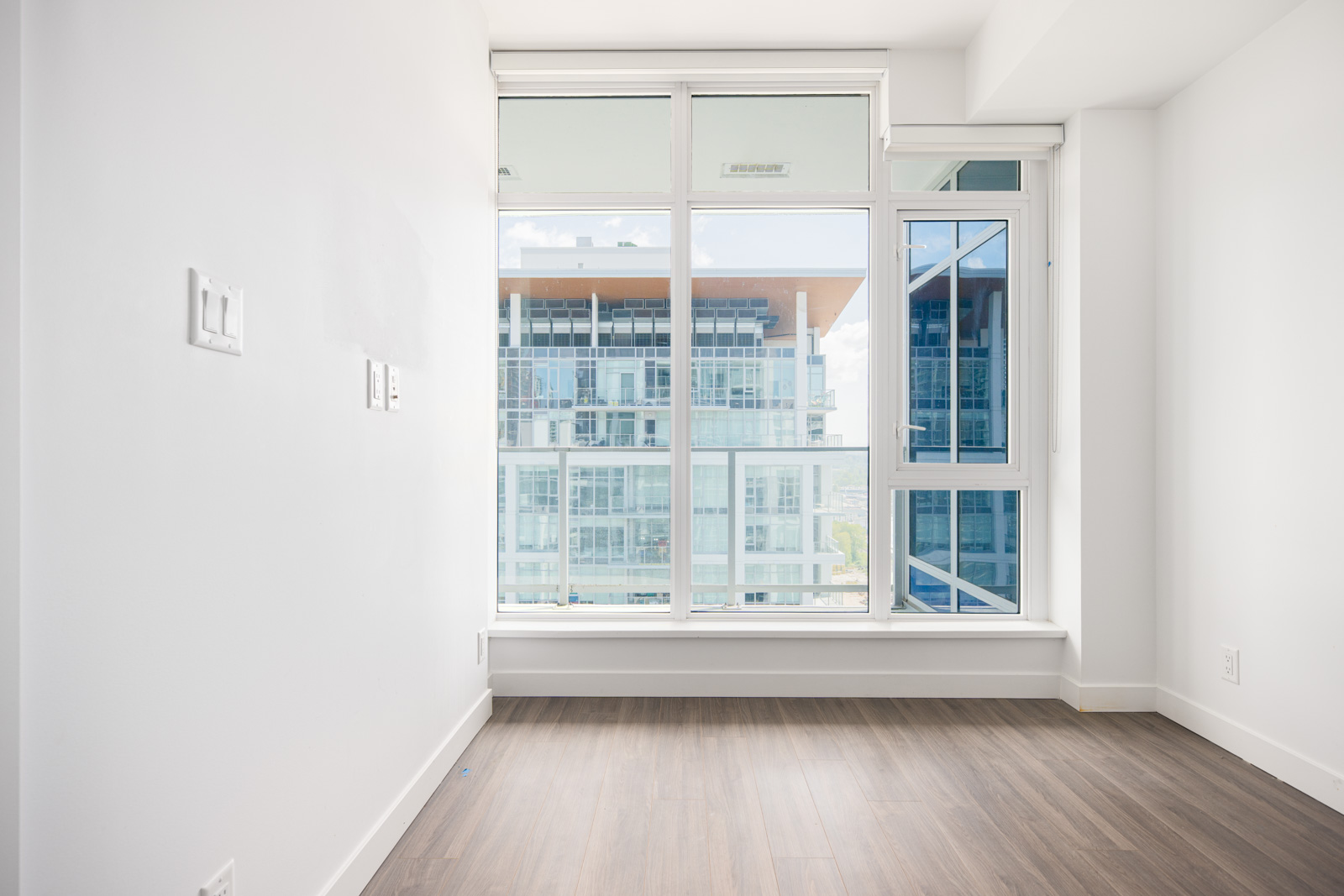 Empty room with white walls and wood flooring featuring large floor-to-ceiling windows overlooking a modern apartment building.