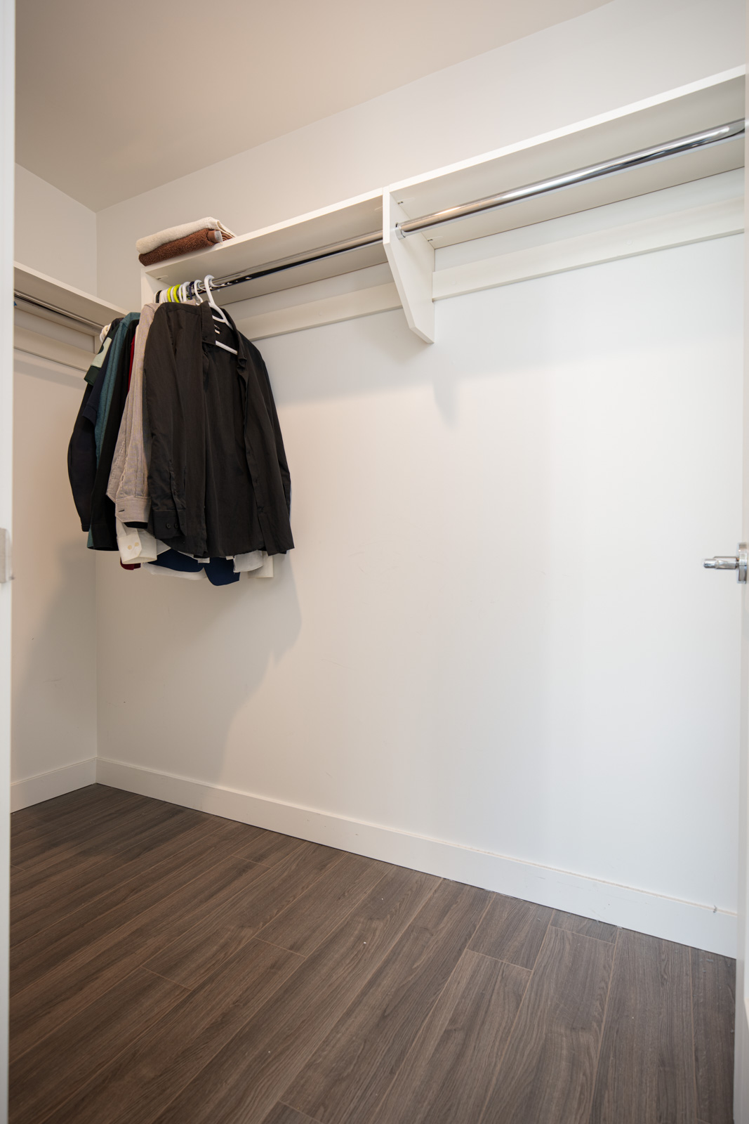 A mostly empty walk-in closet with a few jackets and shirts hanging on a rack, a folded towel on the shelf, and dark wood flooring.