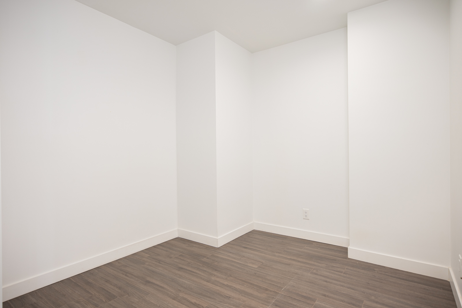 Empty white room with plain walls, gray wood-style flooring, and a visible electrical outlet; no furniture or decorations present.