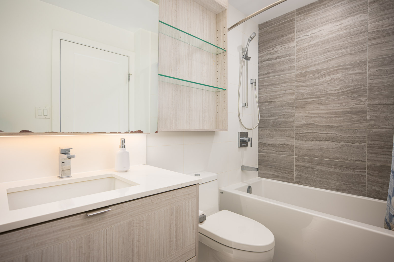 Modern bathroom with a light wood vanity, white countertop, mounted sink, wall-mounted shelves, and a bathtub with a gray tiled shower wall.