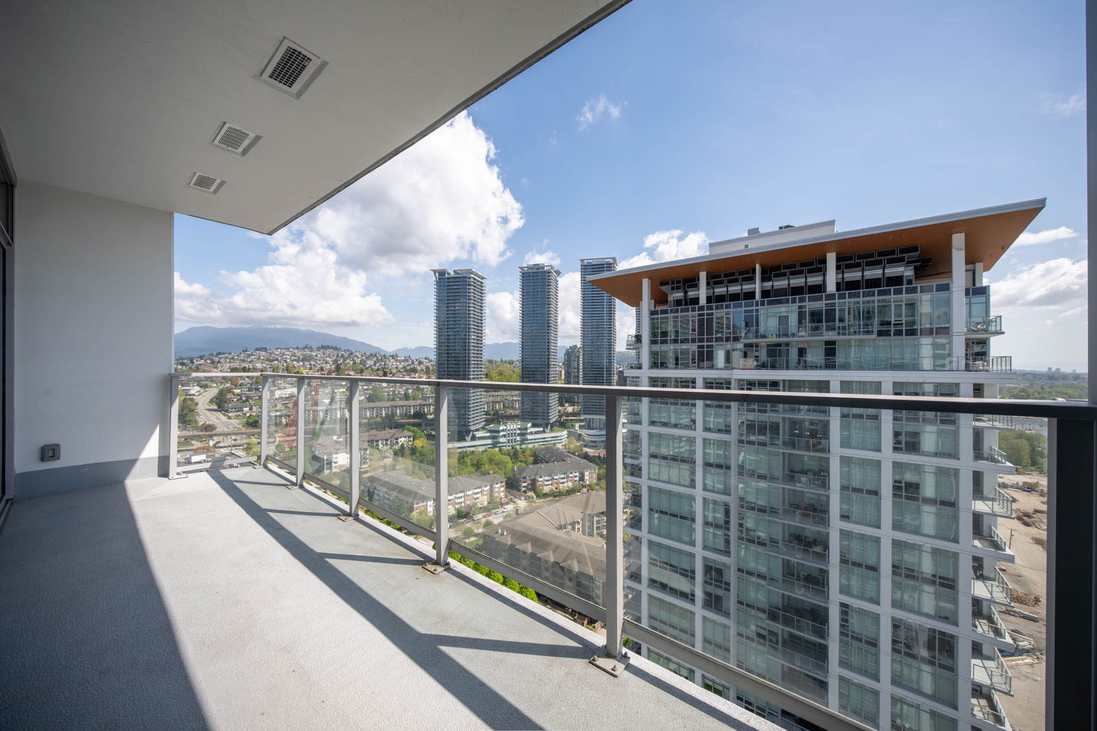 View from a high-rise balcony overlooking a cityscape with glass buildings, distant mountains, and a partly cloudy sky.