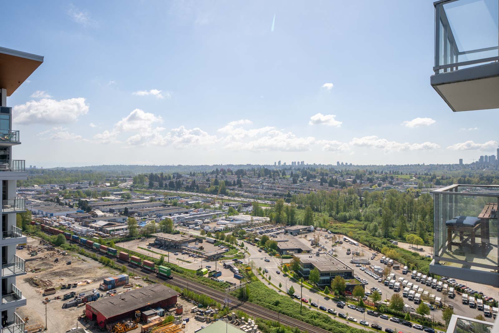 A high-angle view of a suburban area with buildings, roads, railway tracks, parked vehicles, greenery, and a clear sky with scattered clouds.
