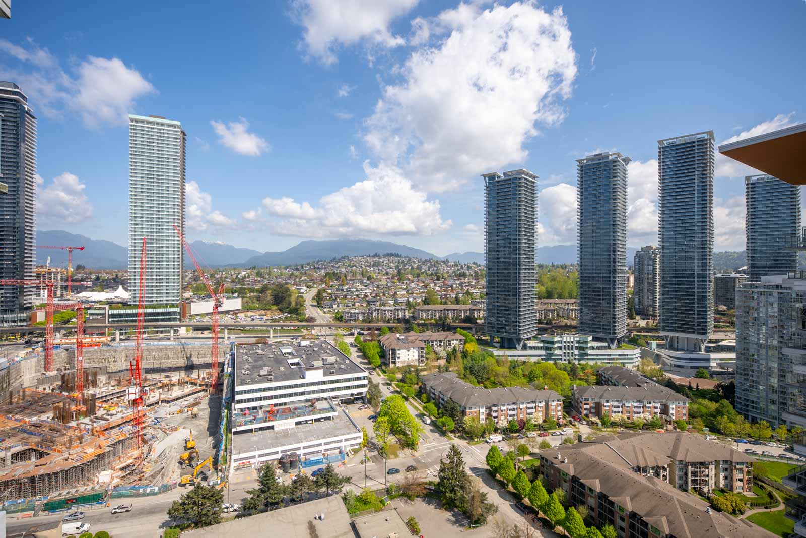 A cityscape with tall modern buildings, construction sites with cranes, residential complexes, and mountains in the background under a partly cloudy sky.