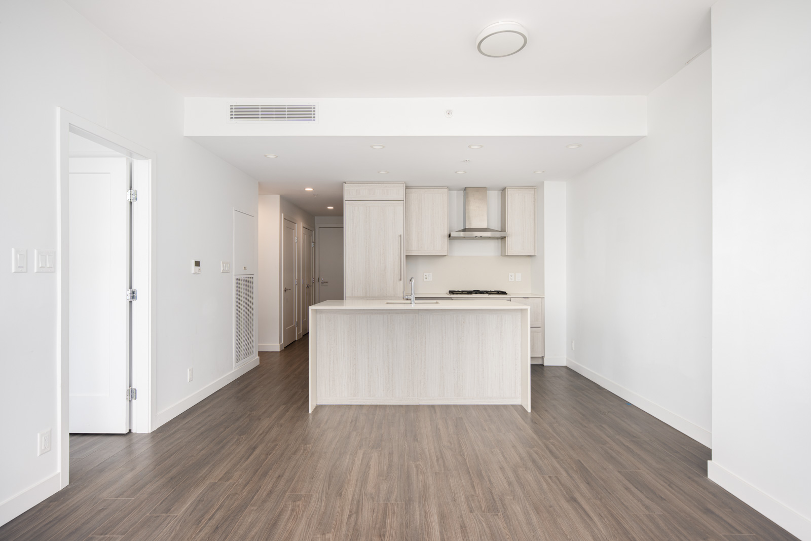 Modern kitchen with light wood cabinets, an island with sink, and stainless steel appliances, set in an open, unfurnished space with dark wood flooring and white walls.