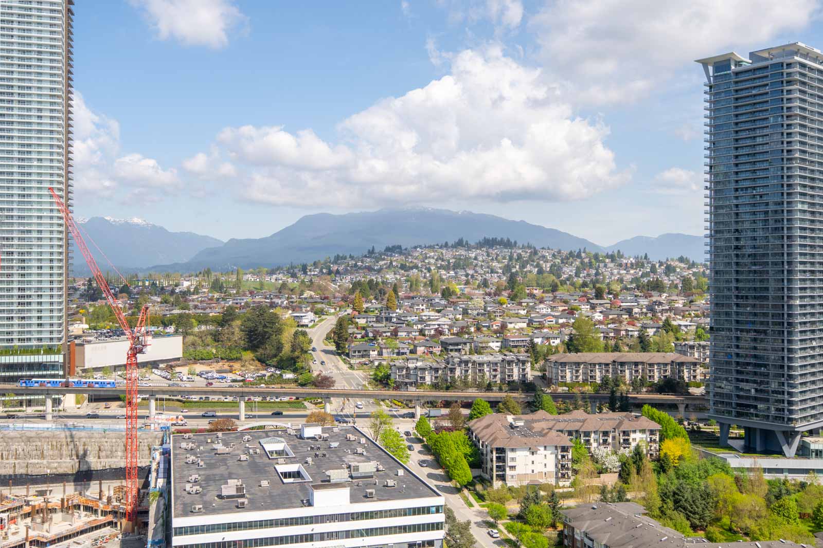 A cityscape featuring high-rise buildings, residential houses, a construction crane, and mountains in the background under a partly cloudy sky.
