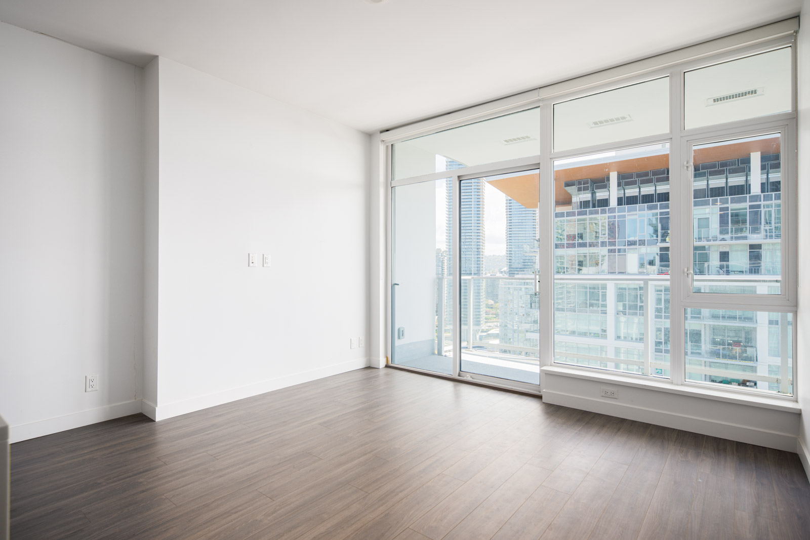 Empty modern apartment room with white walls, large windows, sliding glass door to balcony, and wood flooring; city buildings visible outside.