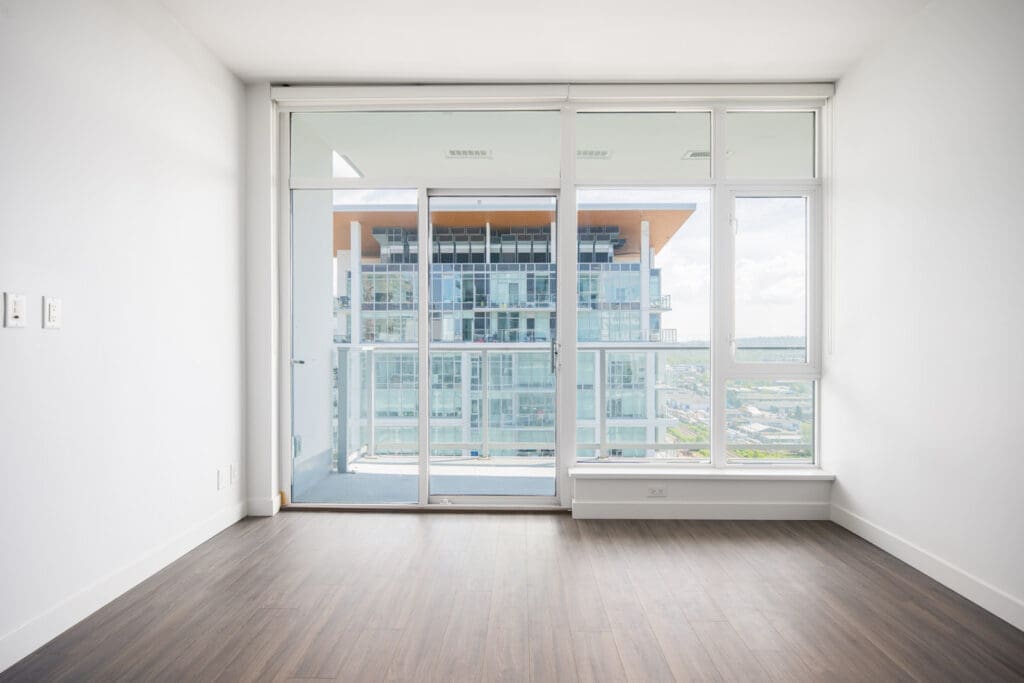 Empty room with white walls, wood flooring, and large glass doors leading to a balcony with a view of a modern building outside.