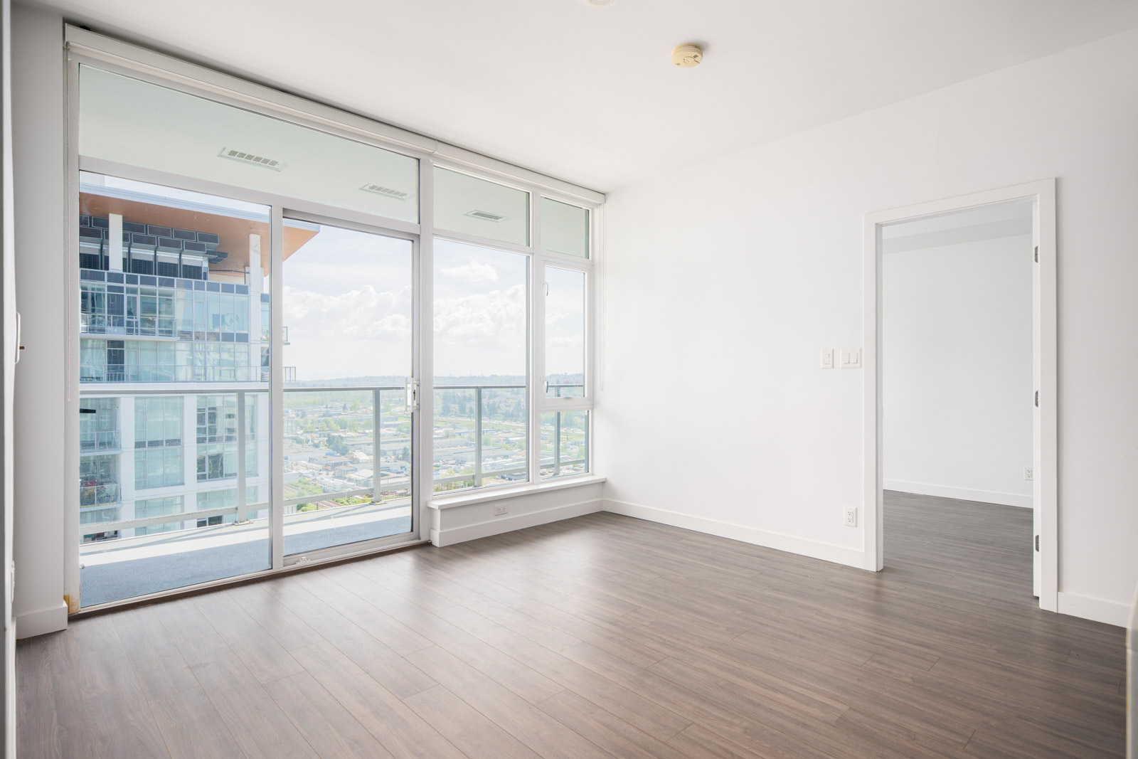Bright, empty modern apartment with large floor-to-ceiling windows, light-colored walls, wood flooring, and a view of other buildings and the cityscape outside.