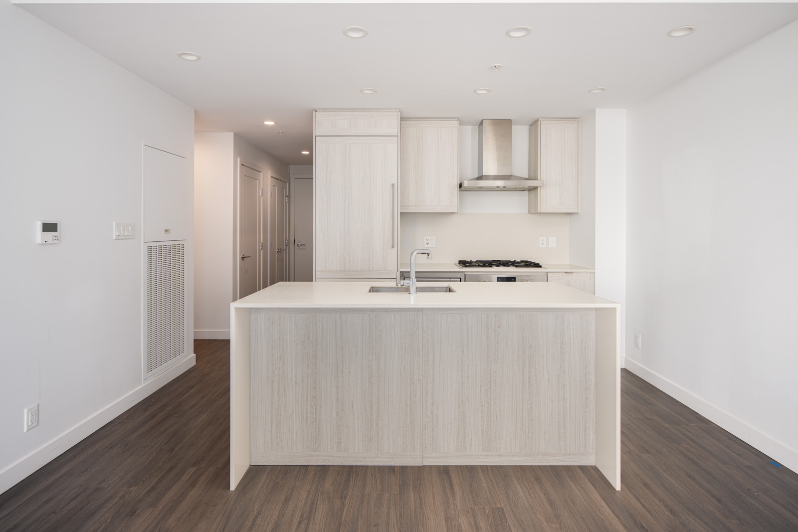 Modern kitchen with light wood cabinets, center island with sink, built-in oven and stove, stainless steel range hood, and dark wood flooring.