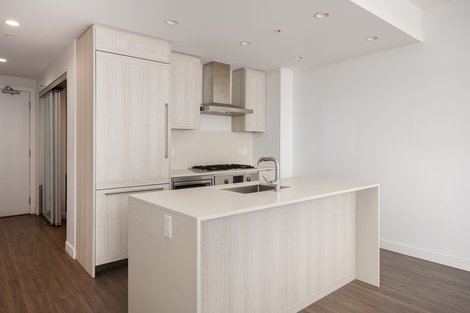 Modern kitchen with light wood cabinets, built-in appliances, a gas stove, stainless steel range hood, and a central island with a sink, set against white walls and a wood floor.
