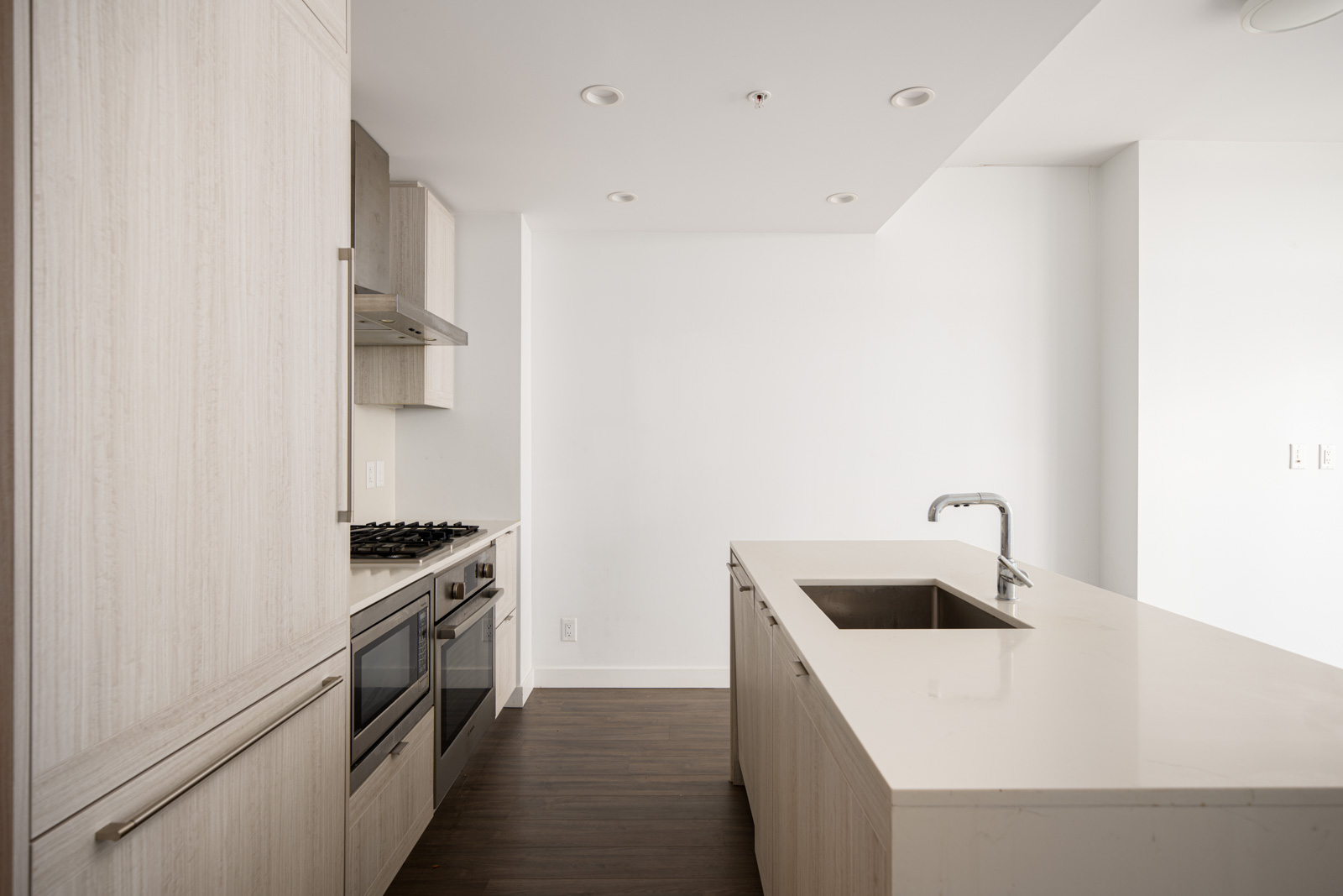 Modern kitchen with light wood cabinets, built-in oven and stove, white countertops, island with sink, and dark wood flooring.