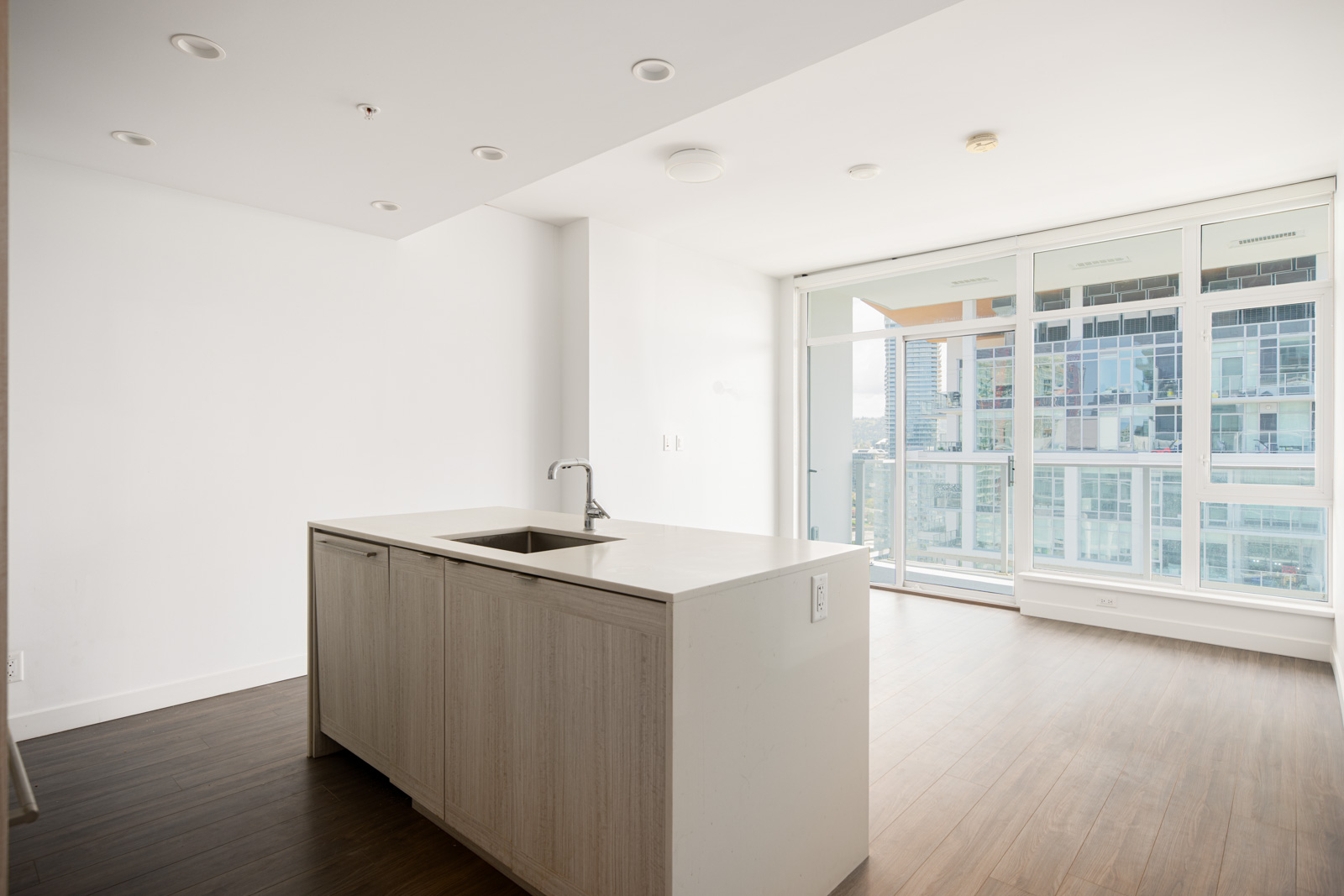 Modern, unfurnished apartment with an open kitchen featuring a central island, large windows, and wood flooring, overlooking a cityscape.
