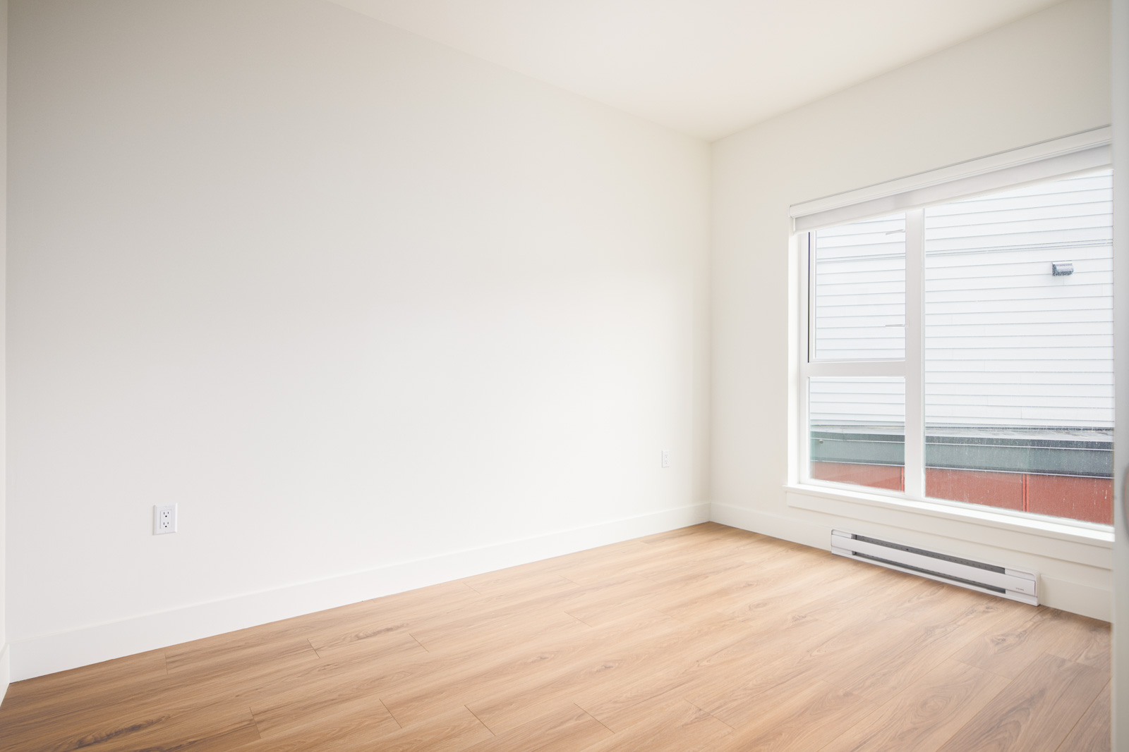 Empty room with white walls, wood-style flooring, a large window with blinds, and a baseboard heater below the window.