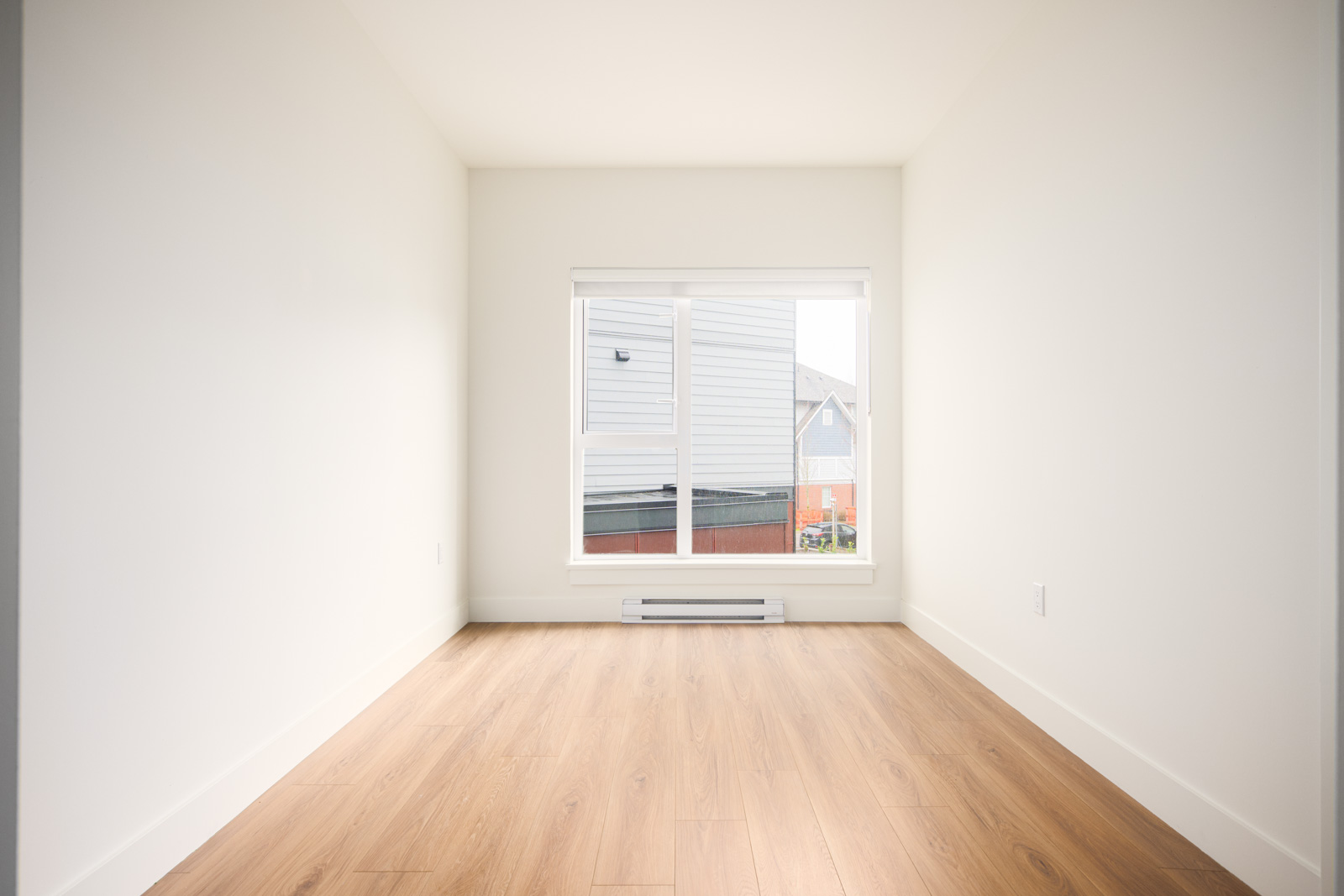 Empty room with white walls, light wood flooring, a large window, and a baseboard heater below the window.