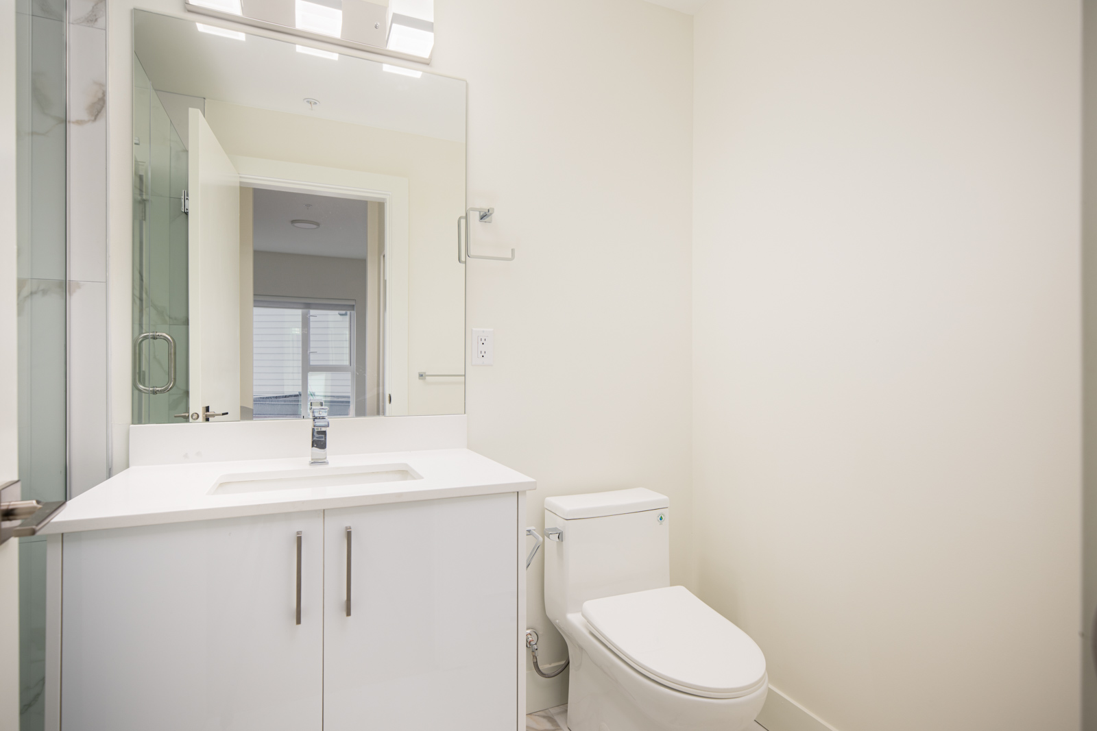 Modern bathroom with a white vanity, large mirror, toilet, and neutral walls; door is open, letting in natural light.