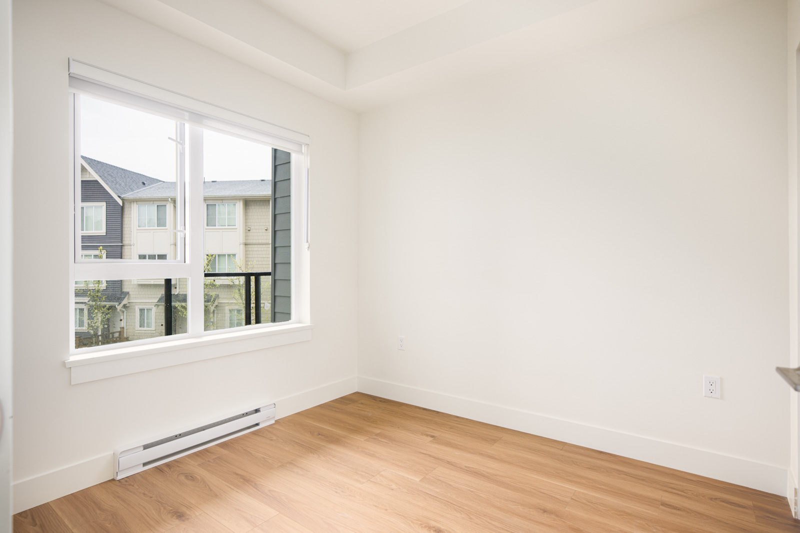 Empty room with light-colored walls, a large window with a view of neighboring buildings, and wood flooring. Natural daylight fills the space.
