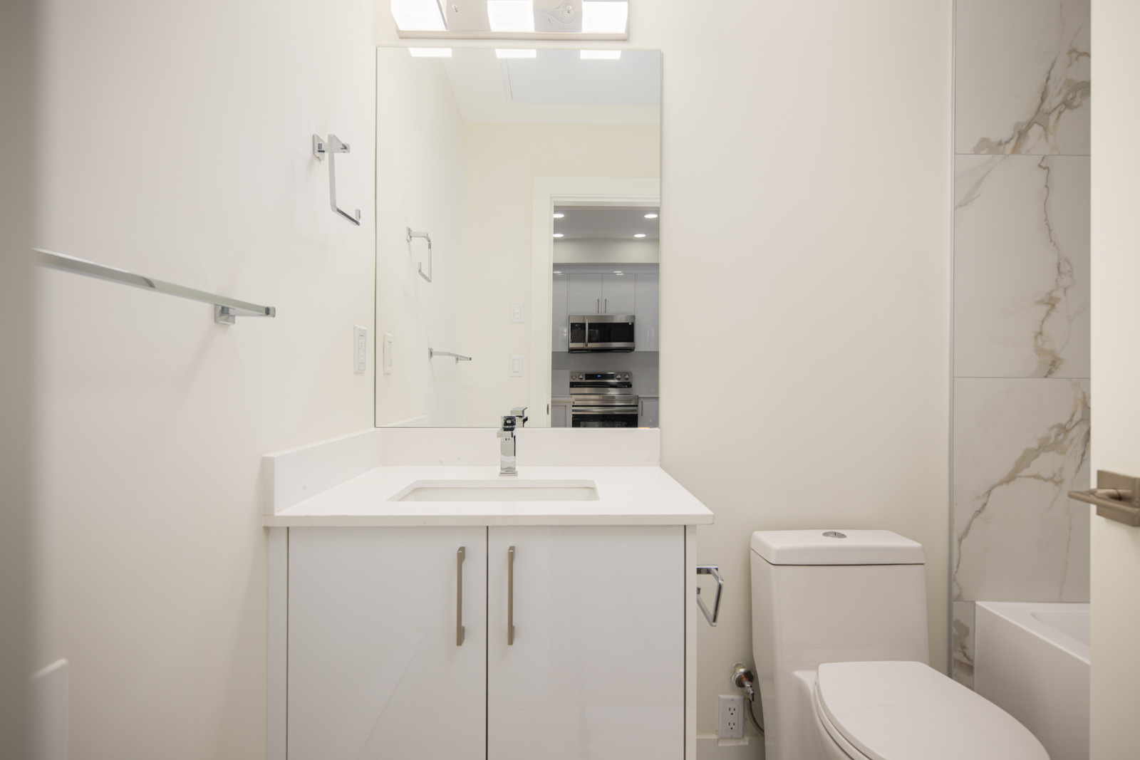 Modern bathroom with a white vanity, rectangular sink, wall-mounted mirror, toilet, and partial view of a shower with marble-style tiles. Kitchen visible in the mirror’s reflection.