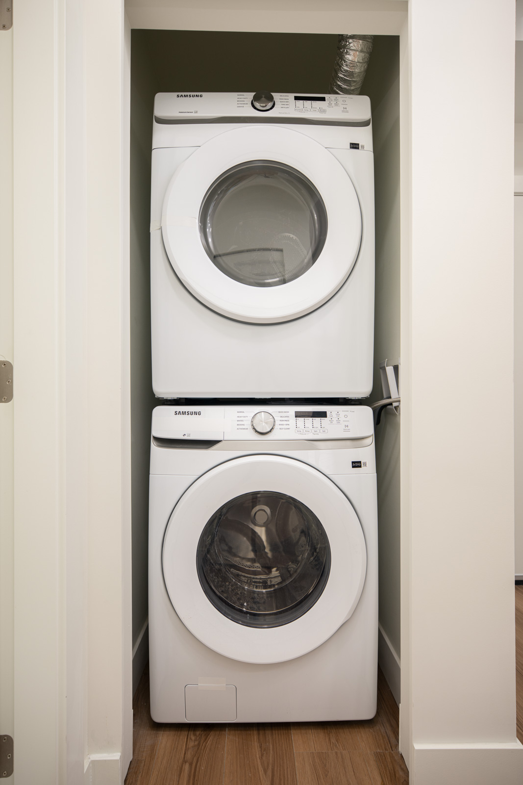 A stacked washer and dryer unit is installed in a small, closet-style laundry space with white walls and a wood floor.