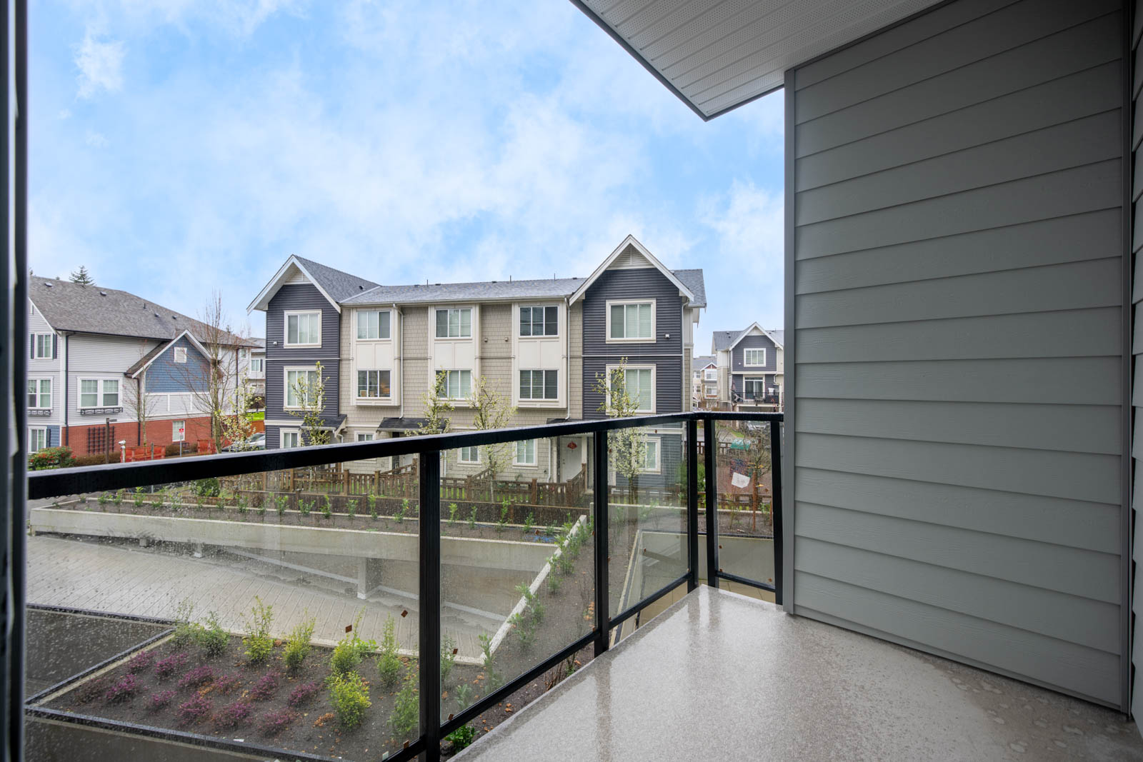 View from a covered balcony with glass railing, overlooking a landscaped area and neighboring multi-story residential buildings on a cloudy day.