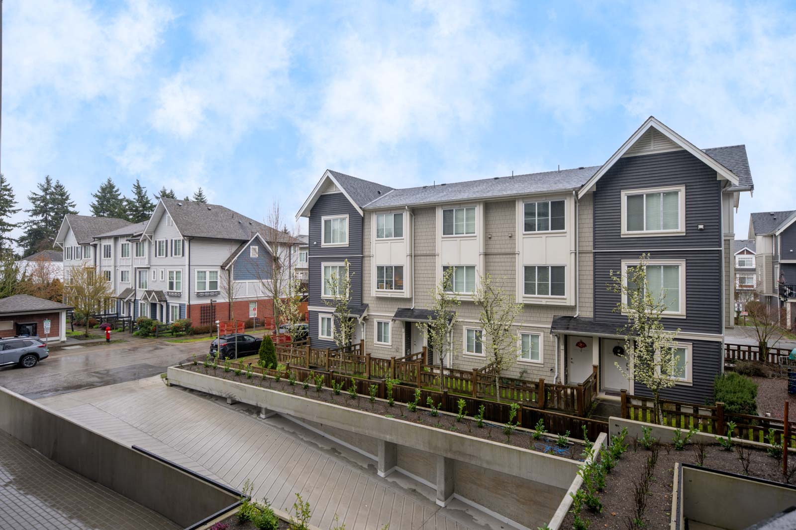 A row of modern, multi-story townhouses with fenced yards and a driveway, under a partly cloudy sky.