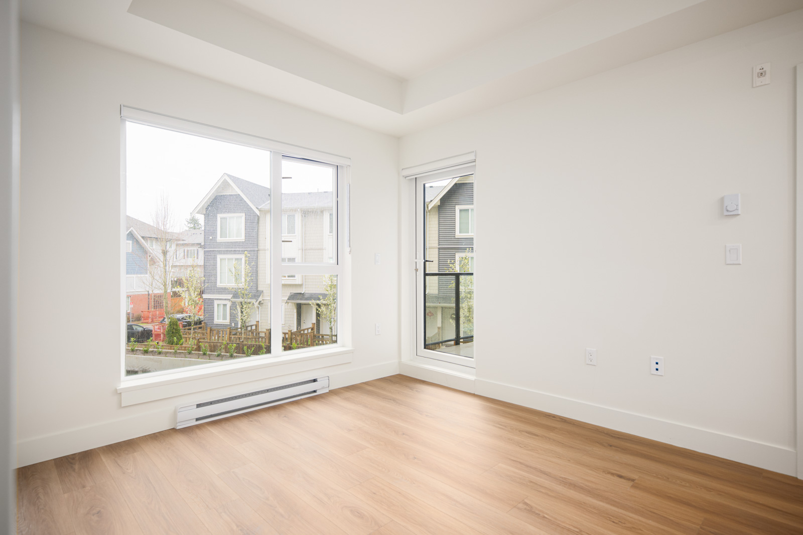 Empty room with light-colored walls, large windows, wood flooring, and a view of neighboring houses outside.