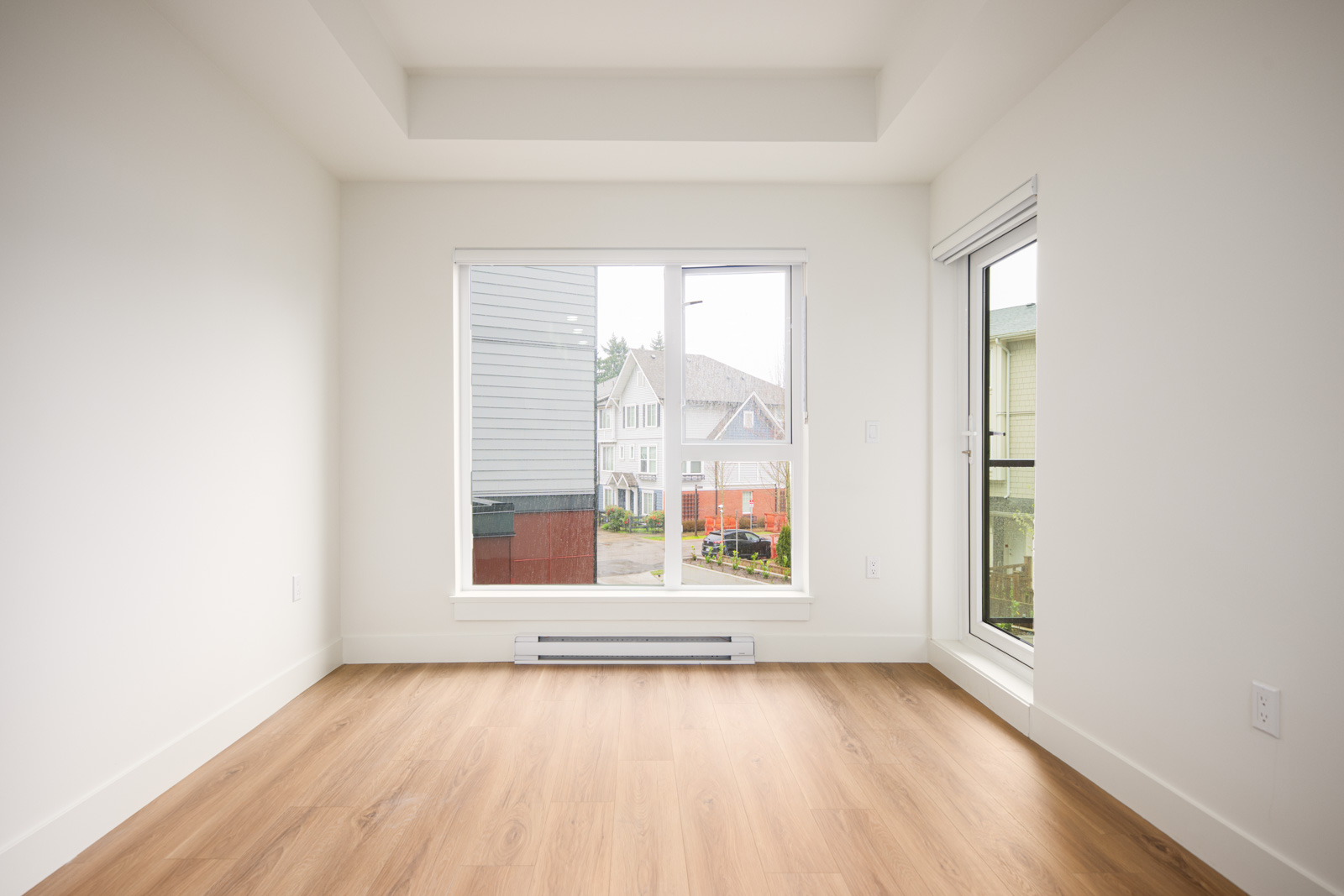 Unfurnished room with light wood flooring, white walls, large window and glass door, overlooking a residential street with houses.