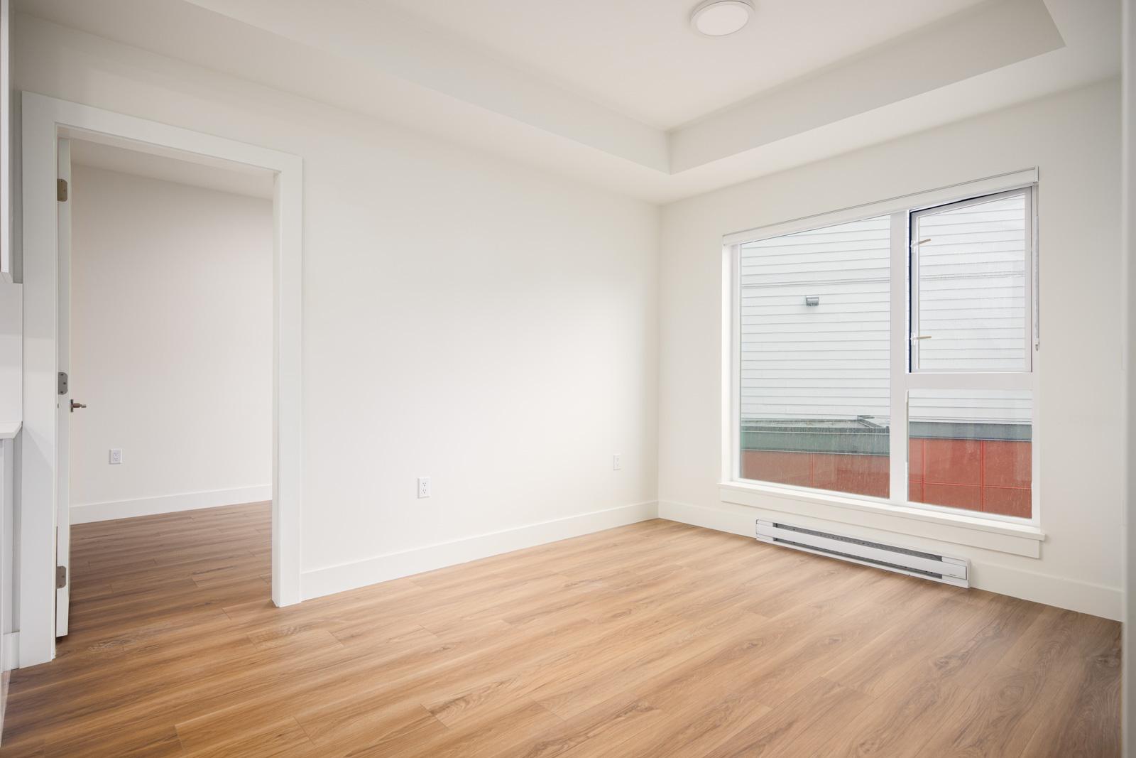 Empty room with white walls, a large window, wood-style flooring, and an open doorway leading to another room. Natural light enters through the window on the right.