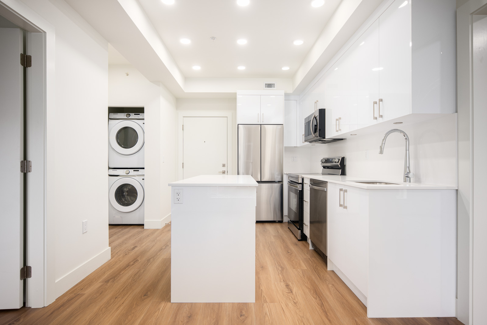 Modern white kitchen with stainless steel appliances, an island, wood flooring, and a stacked washer and dryer unit in a nearby alcove.
